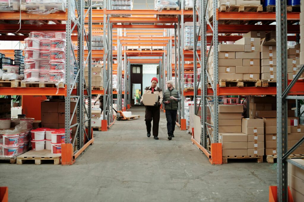 pexels photo 4487383 4487383 Two workers handle a package in a spacious warehouse surrounded by shelves stocked with boxes and products.